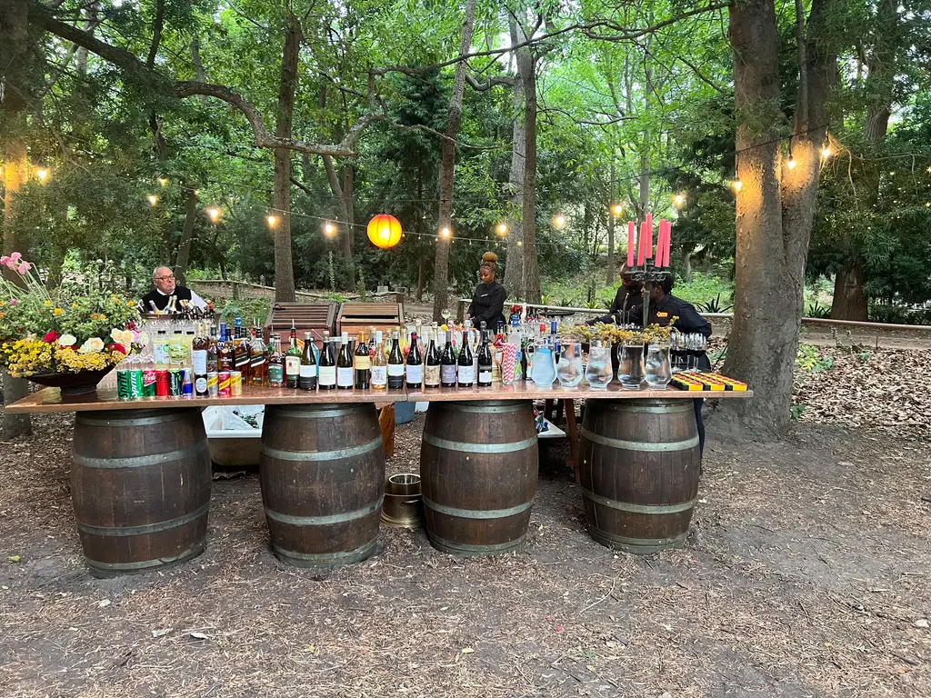 A wine-barrel bar set out in a forest clearing with string lights, flowers and a full line of bottles