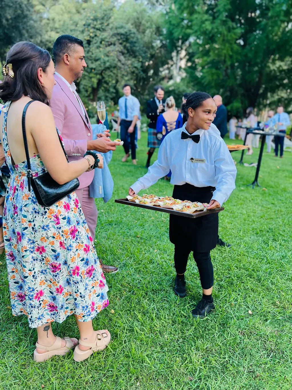 A waitress in a white shirt and bow tie offering canapés to guests on the lawn
